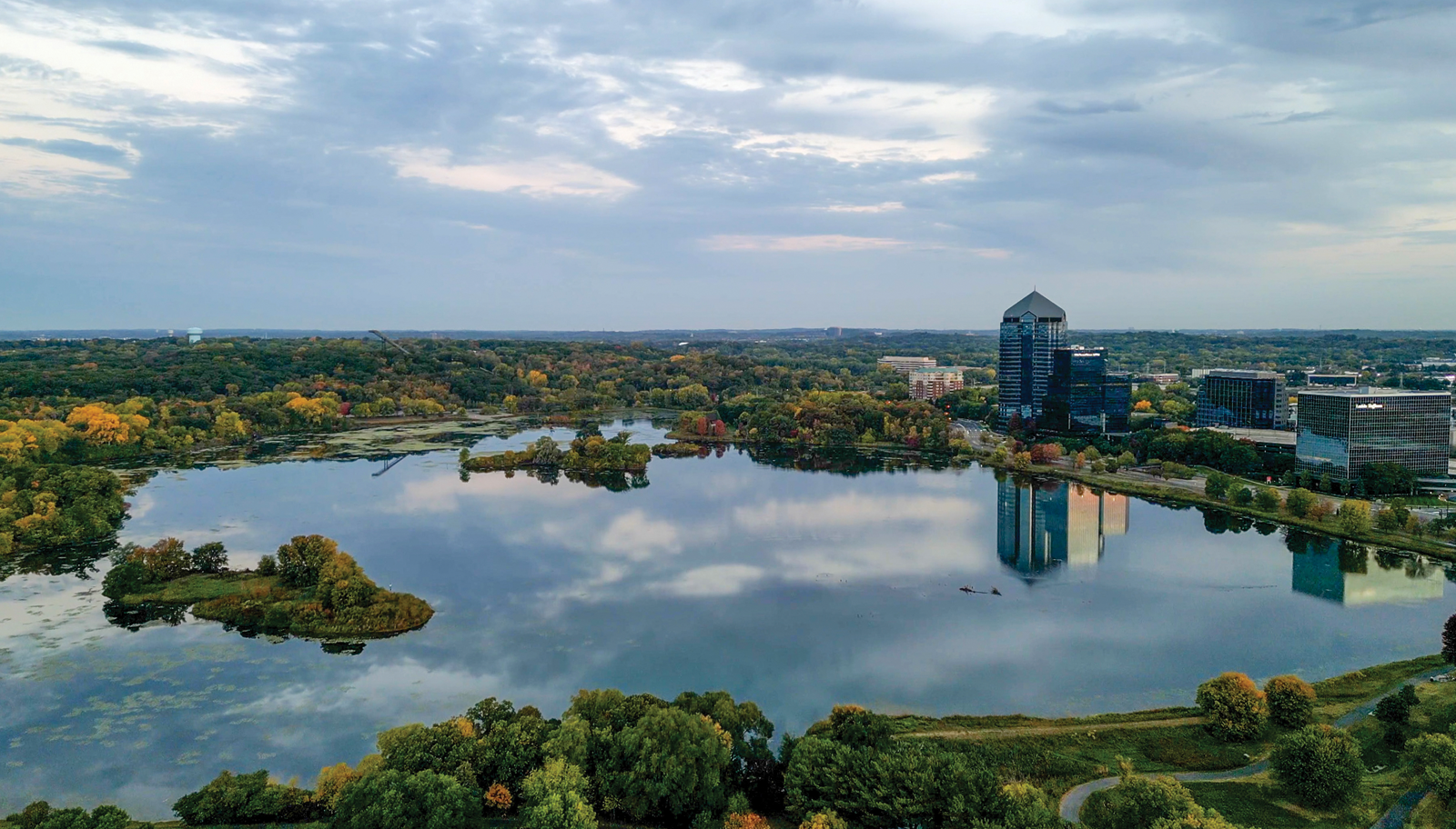 An aerial view of Normandale Lake Park, with greenery and trails around a lake, all below a cloudy sky. Glassy office towers are visible in the background, with their reflections visible in the lake.